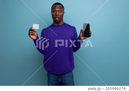 handsome young african guy in blue hoodie uses phone and bank card handsome young african guy in blue hoodie uses phone and bank card 105990274