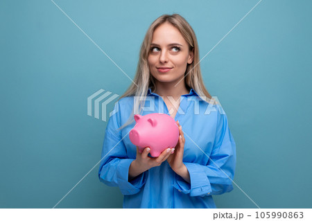 pensive blond girl with an idea holding a piggy bank on a blue isolated background pensive blond girl with an idea holding a piggy bank on a blue isolated background 105990863