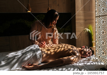 Side view of professional female massage therapist giving massage to handsome man in luxury spa salon lying closed eyes on massage table in sunlight and beautiful shadows. Concept of healthy lifestyle 105991757