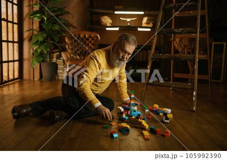 Senior old man playing toy building blocks while sitting on home floor 105992390