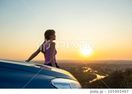 Young woman driver standing near her car enjoying warm sunset view. Girl traveler leaning on vehicle hood looking at evening horizon. Young woman driver standing near her car enjoying warm sunset view. Girl traveler leaning on vehicle hood looking at evening horizon. 105993407
