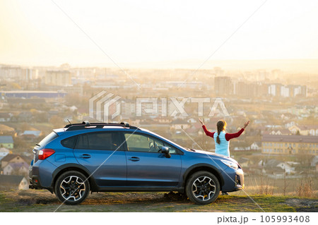 Young woman driver standing beside her offroad car enjoying view of distant city in foggy valley. Travelling and vacation concept Young woman driver standing beside her offroad car enjoying view of distant city in foggy valley. Travelling and vacation concept 105993408