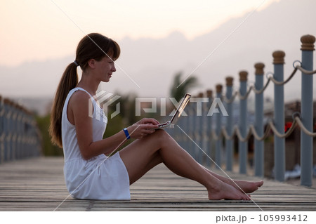 Young woman browsing internet on laptop computer outdoors on warm summer evening. Remote work and study concept. Young woman browsing internet on laptop computer outdoors on warm summer evening. Remote work and study concept. 105993412
