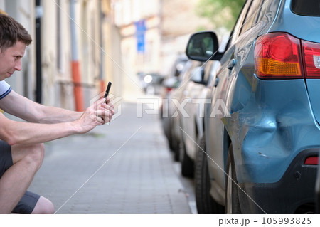 Stressed driver taking picture on sellphone camera of smashed vehicle parked on street side calling for emergency service help after car accident. Road safety and insurance concept Stressed driver taking picture on sellphone camera of smashed vehicle parked on street side calling for emergency service help after car accident. Road safety and insurance concept 105993825