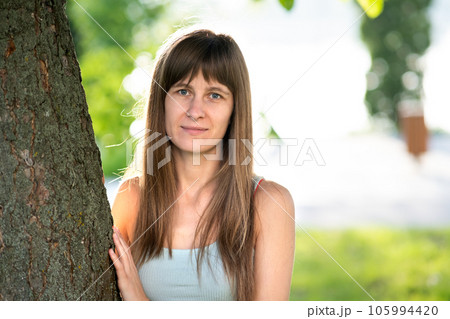 Portrait of pretty young woman with long hair standing neat tree trunk in summer. 105994420