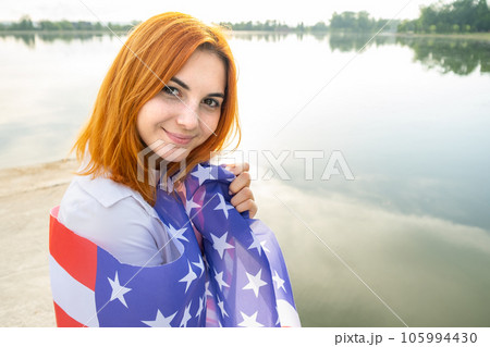 Portrait of happy smiling red haired girl with USA national flag on her shoulders. Young woman celebrating United States independence day. Portrait of happy smiling red haired girl with USA national flag on her shoulders. Young woman celebrating United States independence day. 105994430