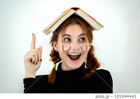 Portrait of funny young smiling student girl with an open book on her head holding her point finger up having an idea. Reading and education concept. Portrait of funny young smiling student girl with an open book on her head holding her point finger up having an idea. Reading and education concept. 105994443