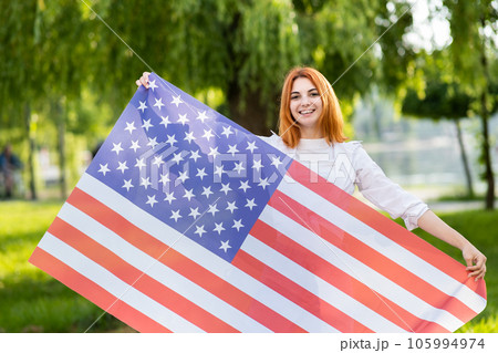 Happy young red haired woman posing with USA national flag standing outdoors in summer park. Positive girl celebrating United States independence day. Happy young red haired woman posing with USA national flag standing outdoors in summer park. Positive girl celebrating United States independence day. 105994974