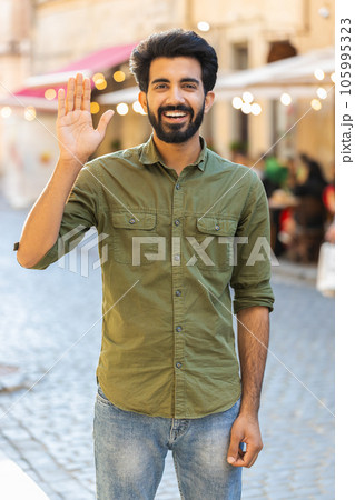 Young man smiling friendly at camera, waving hands hello, hi, greeting or goodbye in city street 105995323