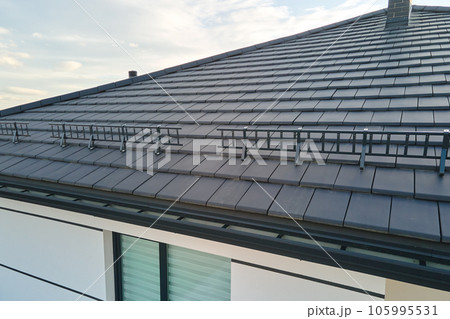 Closeup snow guard for safety in winter on house roof top covered with ceramic shingles. Tiled covering of building Closeup snow guard for safety in winter on house roof top covered with ceramic shingles. Tiled covering of building 105995531