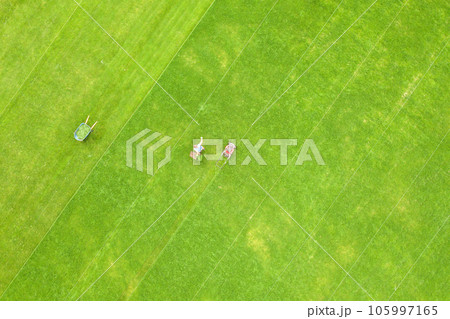 Aerial view of small figure of man worker trimming green grass with mowing mashine on football stadium field in summer. 105997165