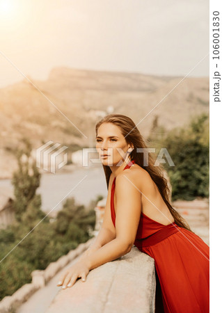 Woman red dress. Summer lifestyle of a happy woman posing near a fence with balusters over the sea. Woman red dress. Summer lifestyle of a happy woman posing near a fence with balusters over the sea. 106001830