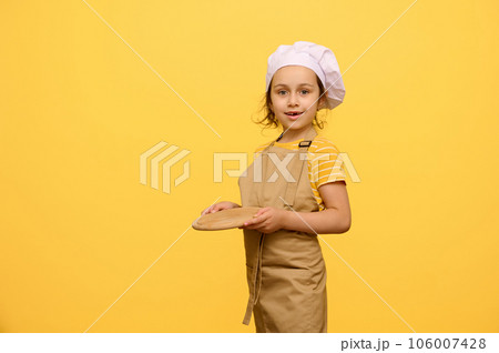 Adorable kid schoolgirl dressed as chef confectioner, holds a wooden board, smiles looking at camera, isolated on yellow Adorable kid schoolgirl dressed as chef confectioner, holds a wooden board, smiles looking at camera, isolated on yellow 106007428