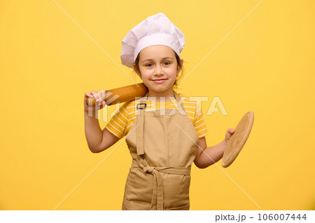 Adorable girl in chef hat and apron, smiles, looking at camera, holding rolling pin and wooden board, isolated on yellow Adorable girl in chef hat and apron, smiles, looking at camera, holding rolling pin and wooden board, isolated on yellow 106007444