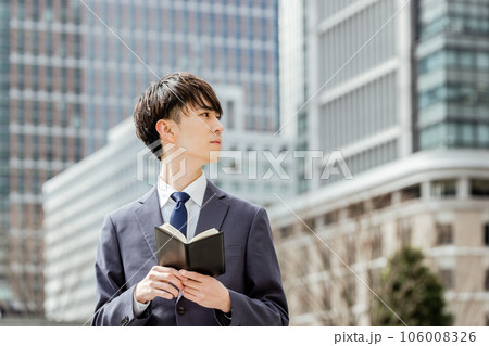 建物に囲まれたビジネスオフィス街で真剣な表情で手帳を広げながら遠くを見つめる20代のスーツの男 106008326