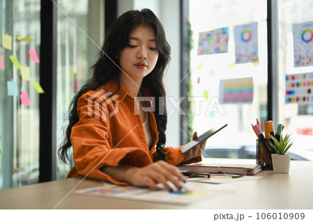 Beautiful stylish Asian woman in orange jacket using digital tablet, working in creative office. 106010909