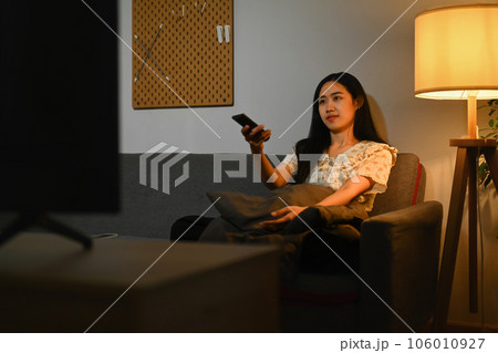 Beautiful asian woman watching television sitting on a comfortable sofa with the remote control in her hand. 106010927