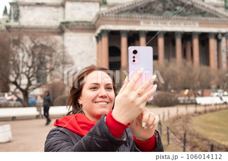 young caucasian woman with smartphone. female making video call, taking selfie portrait with mobile phone on street of European historical city. tourism 106014112
