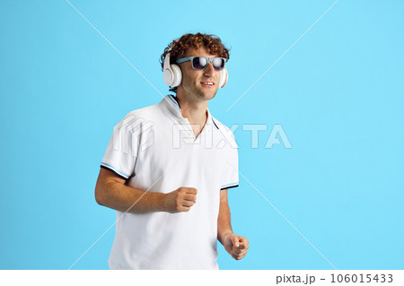 Mature man in casual white t-shirt and sunglasses listening to music in headphone against blue studio background. Enjoyment Mature man in casual white t-shirt and sunglasses listening to music in headphone against blue studio background. Enjoyment 106015433