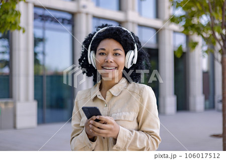 Portrait of young beautiful African American woman, student smiling and looking at camera holding phone in hands using headphones for listening to music and online radio with podcasts. 106017512