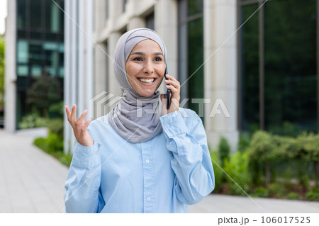 Young joyful businesswoman in hijab talking on the phone outside an office building. 106017525