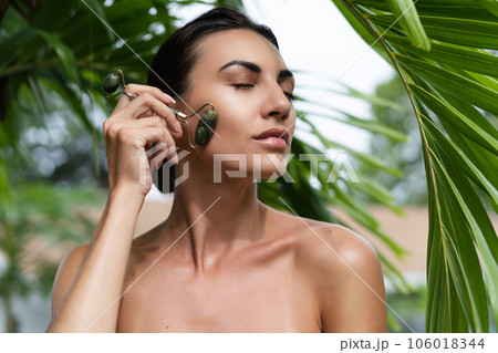 Beauty portrait, soft skin. Female model with natural make-up and healthy skin behind a green leafy plant. Portrait of a beautiful girl with nude nails, bare shoulders holding roller massager outdoor Beauty portrait, soft skin. Female model with natural make-up and healthy skin behind a green leafy plant. Portrait of a beautiful girl with nude nails, bare shoulders holding roller massager outdoor 106018344