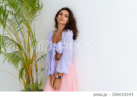 Stylish young woman with voluminous hair in a trendy long sleeve crop top and a pink skirt posing against a white wall and a tropical bush, wearing a smart watch 106018345