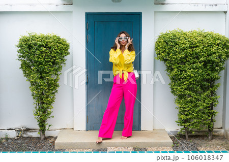Stylish fit fashion women in bright pink wide leg pants and yellow shirt holding bag trendy mint sunglasses posing at luxury tropical villa by pool outdoor natural day light 106018347