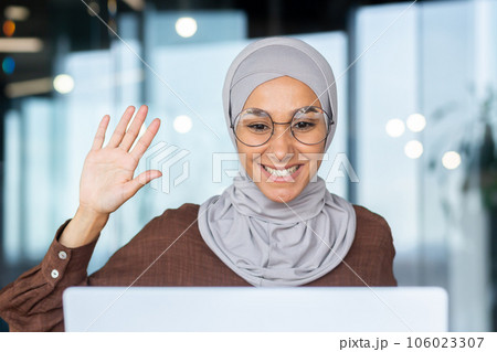Young smiling muslim woman in hijab student talking on video call via laptop. He waves his hand at the camera, says hello. Young smiling muslim woman in hijab student talking on video call via laptop. He waves his hand at the camera, says hello. 106023307