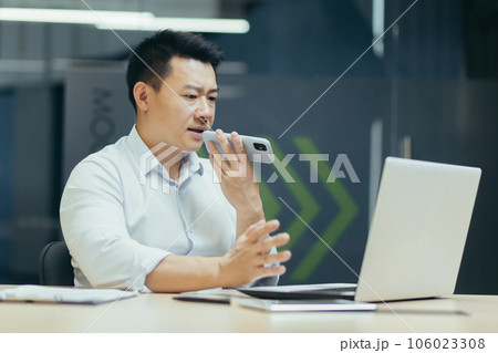 Asian serious young man businessman sitting in office working on laptop and talking on speaker phone, recording conversation. Asian serious young man businessman sitting in office working on laptop and talking on speaker phone, recording conversation. 106023308