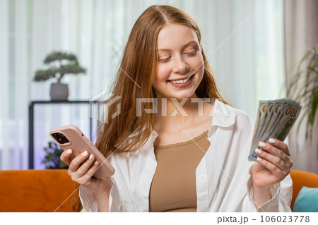 Smiling happy young girl counting money cash use smartphone income saves lottery win budget at home 106023778