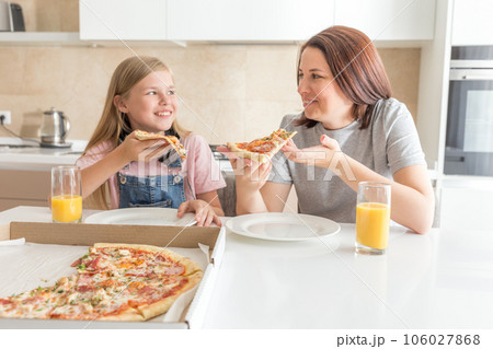 Mother and daughter sitting in the kitchen, eating pizza and having fun. Focus on daughter 106027868