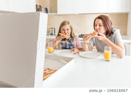 Mother and daughter sitting in the kitchen, eating pizza and having fun. Focus on daughter Mother and daughter sitting in the kitchen, eating pizza and having fun. Focus on daughter 106028067