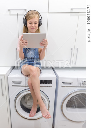 a Happy young girl listens to music on headphones holding tablet in laundry room with washing machine a Happy young girl listens to music on headphones holding tablet in laundry room with washing machine 106028174