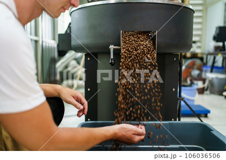 Professional male and female coffee roasters working with modern automated roasting machine in the coffee roasting factory. Coffee roasters checking or inspecting the roasting quality of coffee bean. Professional male and female coffee roasters working with modern automated roasting machine in the coffee roasting factory. Coffee roasters checking or inspecting the roasting quality of coffee bean. 106036506