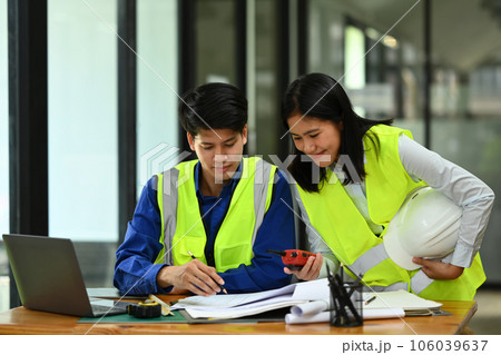 Engineers man and woman wearing yellow vests working with blueprint, discussing the new project at office 106039637