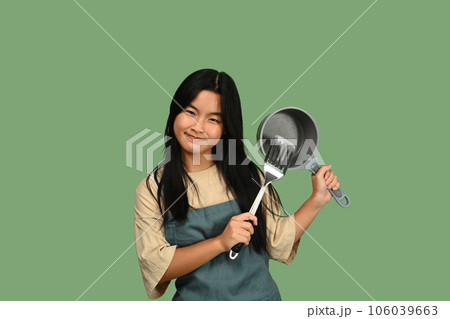 Playful teenage girl wearing apron holding pan and spatula isolated on green background ready for cooking Playful teenage girl wearing apron holding pan and spatula isolated on green background ready for cooking 106039663