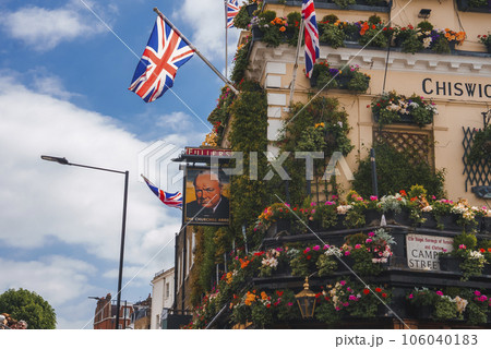 Frame of Winston Churchill on famous pub in city. United Kingdom Flags hanging on pole. Iconic landmark covered with flowers. 106040183