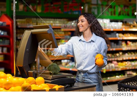 Young woman shopper in supermarket uses self-service scales, smiling Hispanic woman weighs fruits and vegetables in large store. Young woman shopper in supermarket uses self-service scales, smiling Hispanic woman weighs fruits and vegetables in large store. 106043193
