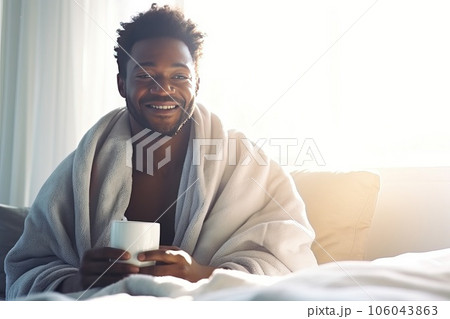 A middle-aged African American sits on a bed with a mug of aromatic coffee. 106043863