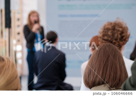 Back view of group of people gathering together in conference hall and listening to presentation of female speaker with microphone Back view of group of people gathering together in conference hall and listening to presentation of female speaker with microphone 106046666