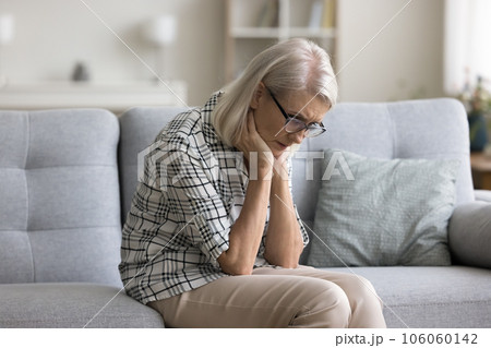 Sad depressed retired blonde woman sitting on sofa at home 106060142