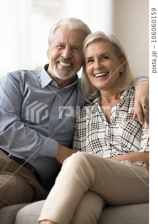 Cheerful positive senior retired husband and wife hugging on sofa 106060159