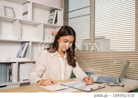 Asian woman working at home, Press the calculator to calculate the income - expenses of the house and use the laptop to record the information and check the accuracy, Home lifestyle. 106062510