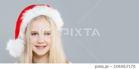 Portrait of Cheerful Christmas Girl in Santa hat smiling and looking at camera on gray background Portrait of Cheerful Christmas Girl in Santa hat smiling and looking at camera on gray background 106063570
