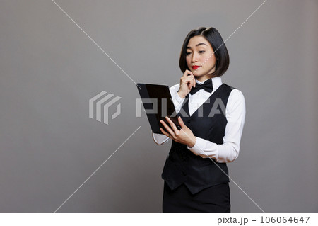 Thoughtful asian waitress in professional uniform managing order on digital tablet. Pensive receptionist thinking while examining restaurant menu on portable gadget and posing in studio Thoughtful asian waitress in professional uniform managing order on digital tablet. Pensive receptionist thinking while examining restaurant menu on portable gadget and posing in studio 106064647