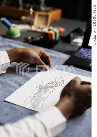 Close up of meticulous couturier drawing stylish dress from upcoming sartorial custom handmade collection in tailoring studio. Tailor creating bespoken sartorial garment piece Close up of meticulous couturier drawing stylish dress from upcoming sartorial custom handmade collection in tailoring studio. Tailor creating bespoken sartorial garment piece 106064835