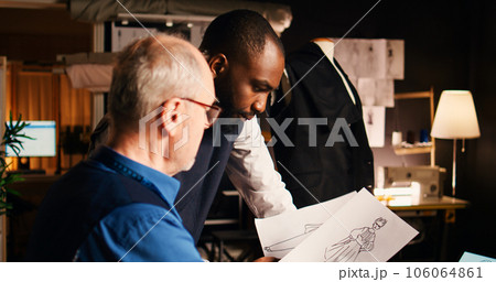 Senior tailor teaching trainee skills to create modern fashion clothing line, tailoring handcraft process. Beginner and experienced designer looking at outlines sketches. Handheld shot. Senior tailor teaching trainee skills to create modern fashion clothing line, tailoring handcraft process. Beginner and experienced designer looking at outlines sketches. Handheld shot. 106064861