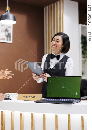 Receptionist having greenscreen on desk, welcoming guests at hotel and starting check in process with tablet. Asian woman using laptop showing isolated display with chroma key copyspace. 106064887