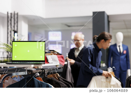 Open laptop computer with chroma key green screen standing on cloth rack in clothing shop, people shopping on background. Small retail business and inventory management software 106064889